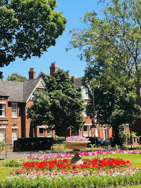 A bright, sunny day in a residential park area with lush green trees and a clear blue sky. In the foreground, there is a vibrant flowerbed featuring red, pink, and white flowers arranged in neat clust
