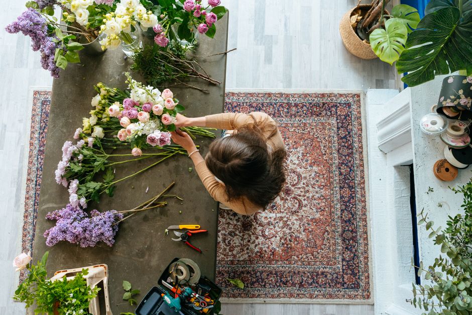 A woman with dark, wavy hair is arranging a floral bouquet on a rustic grey table indoors. The bouquet includes pink and white roses, purple lavender, and greenery. The table has various flower stems