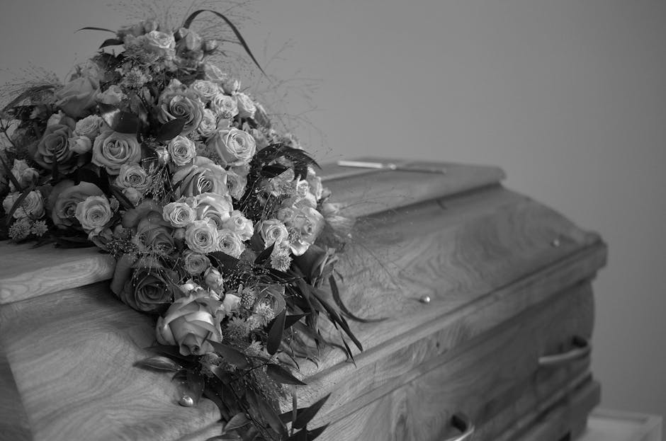 A large, elegant floral bouquet resting on a wooden casket, featuring a mix of roses and smaller blooms with foliage, predominantly in soft pastel shades and darker accents. The arrangement is lush an